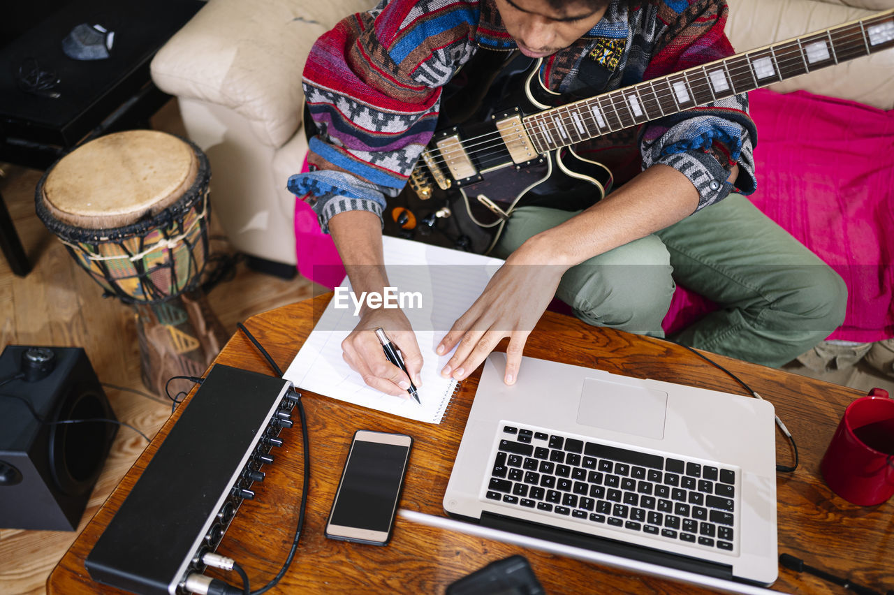 Young man with guitar writing notes on book by laptop in living room