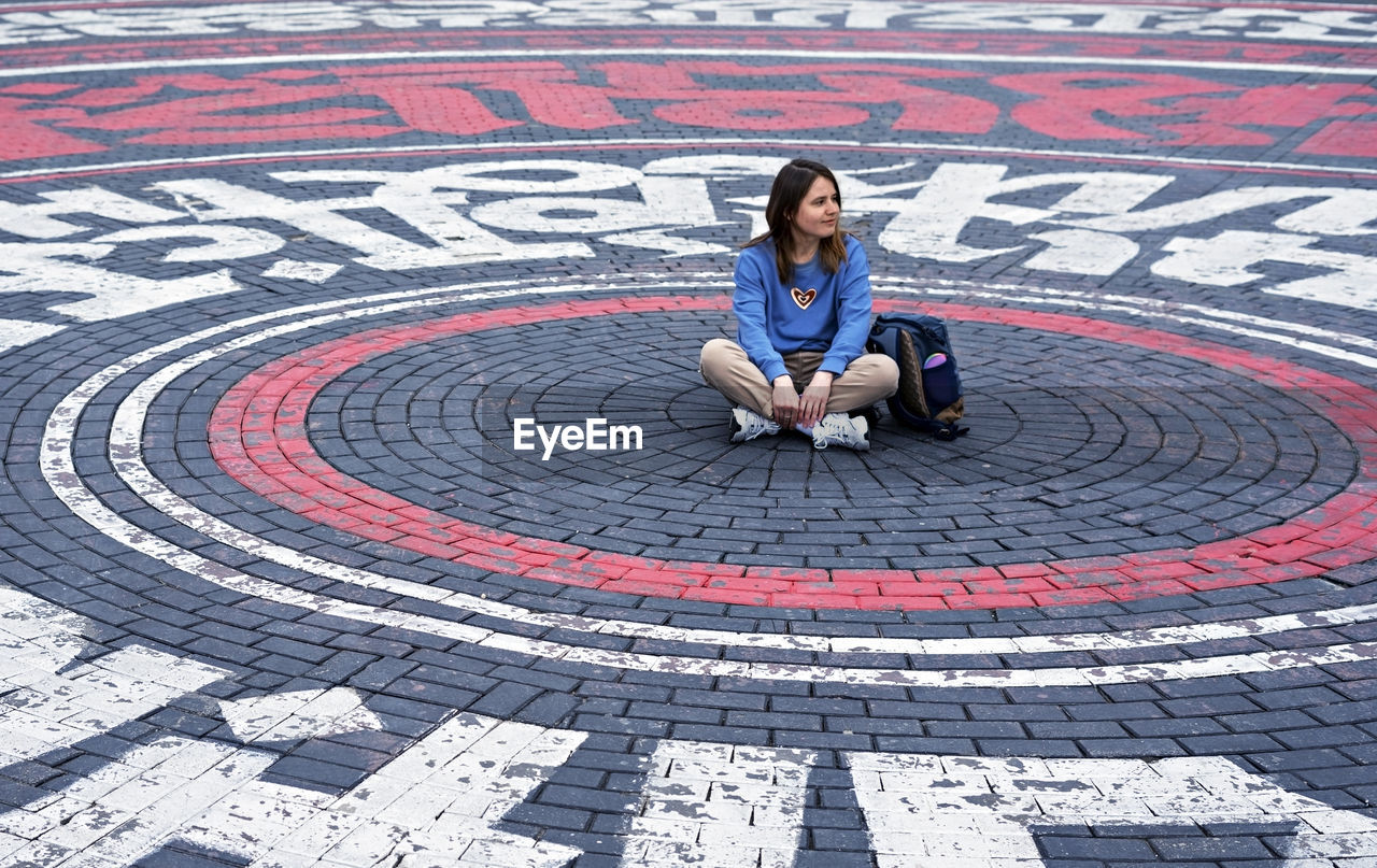 Young woman in blue sweatshirt alone sitting on street cobbled square in the form of circles