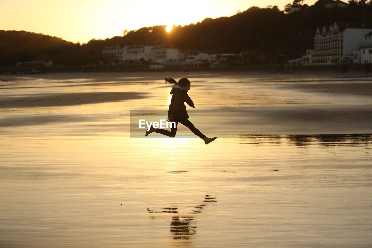 Side view of girl jumping on shore at beach during sunset
