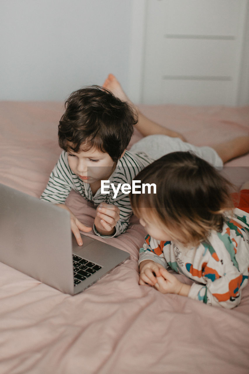 Little brother and sister lie in the bedroom on the bed and look at the laptop. high quality photo