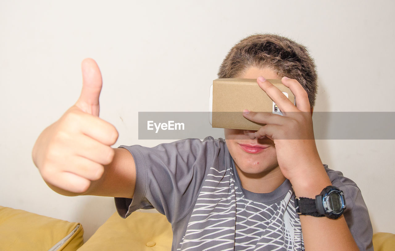 Boy watching virtual reality while gesturing thumbs up sign against gray background