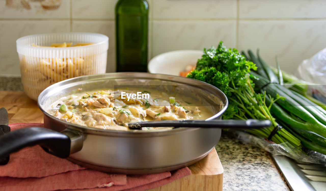 White sauce pasta dish in frying pan on chopping board with parsley, oil, spring onion and pasta