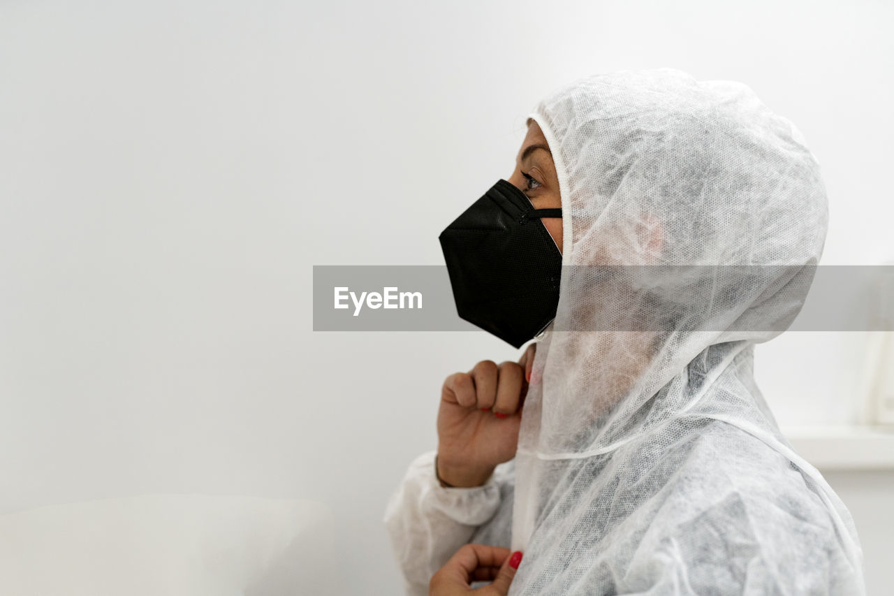 Side view of female medical specialist putting on protective suit with face mask and protective cap while preparing for work during coronavirus pandemic