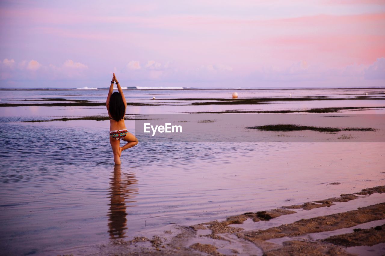 Rear view of bikini woman doing tree pose at beach against sky during sunset