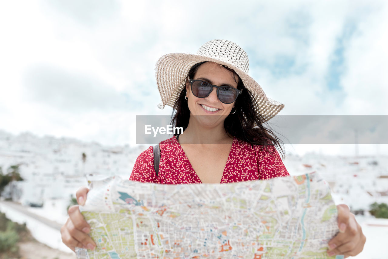 Positive female tourist with map in straw hat and sunglasses standing under cloudy sky during summer trip