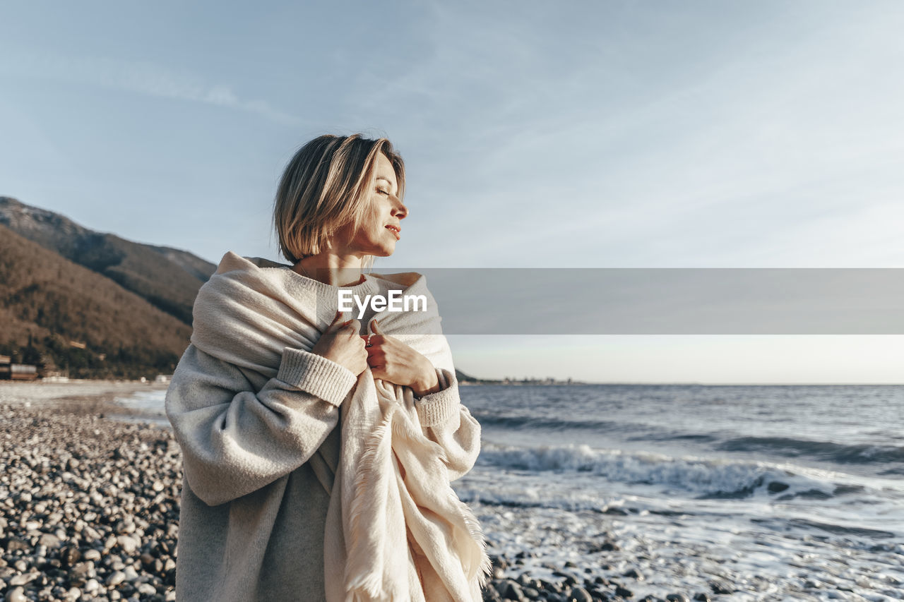 Blond woman wrapped in blanket enjoying sunset at beach