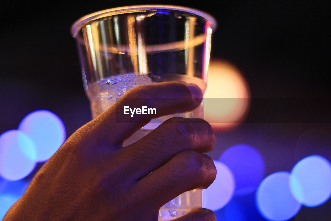 CLOSE-UP OF HAND HOLDING GLASS OF ILLUMINATED BLURRED BACKGROUND