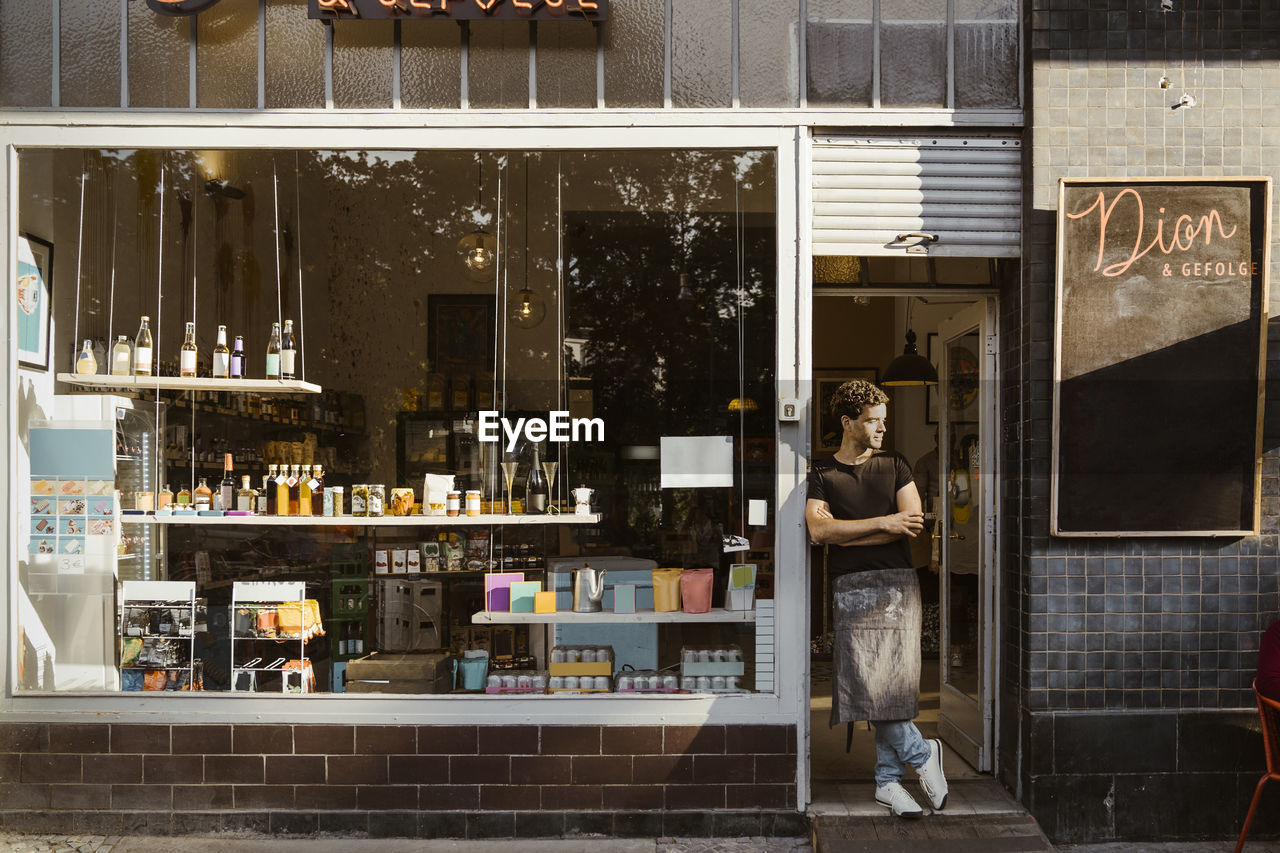 Male shop owner with arms crossed leaning at store doorway