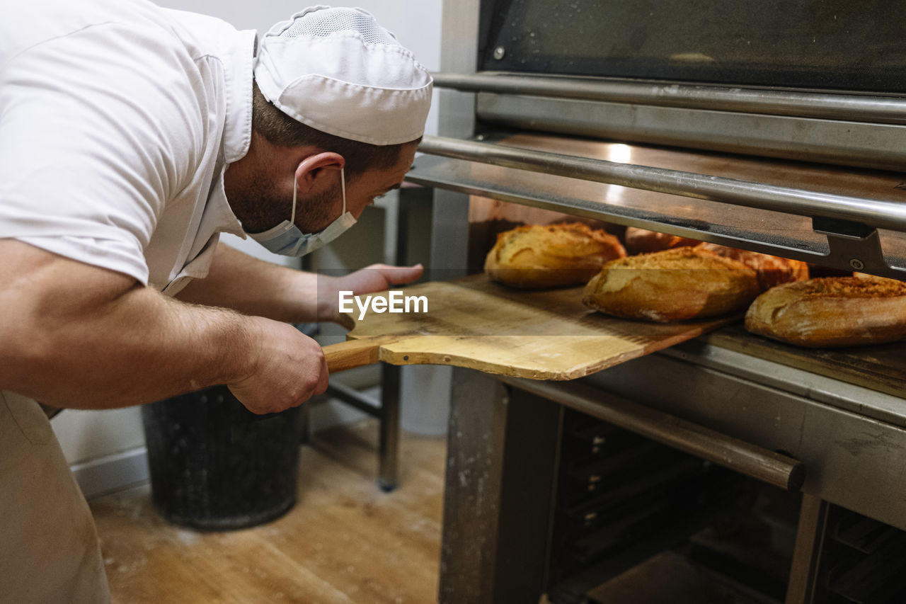 Male baker removing breads from oven in kitchen at bakery