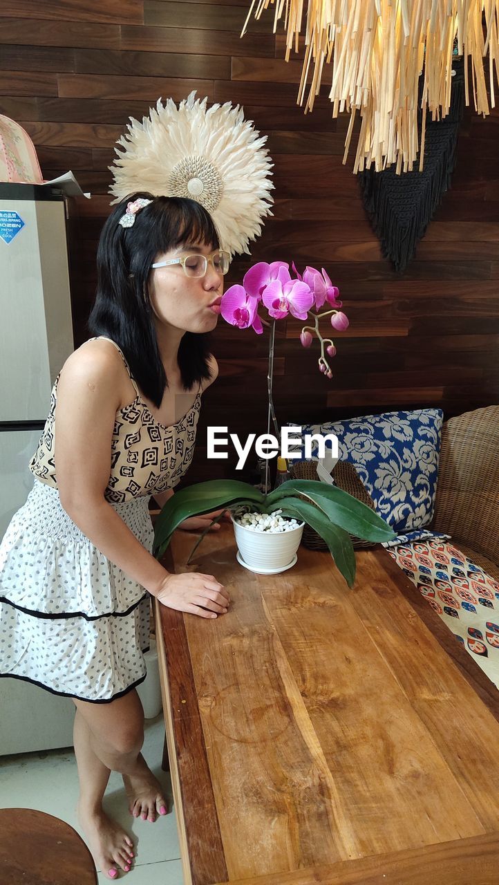 YOUNG WOMAN SITTING ON TABLE BY POTTED PLANTS AT HOME