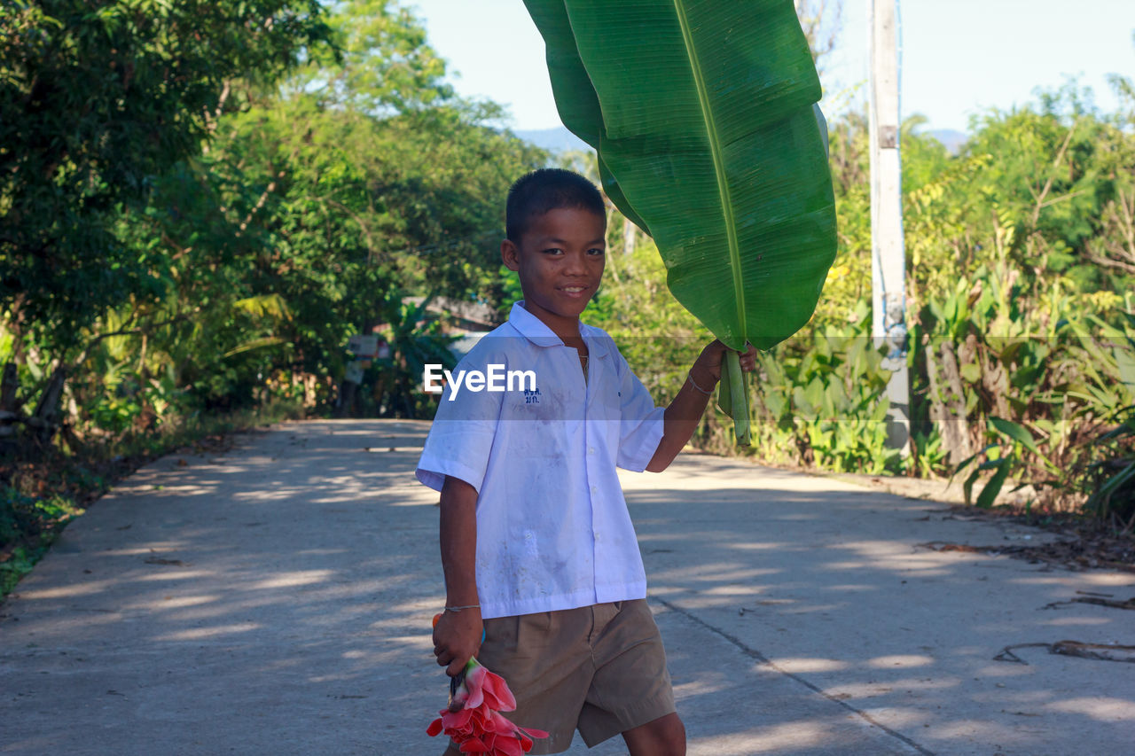 Smiling boy holding banana leaf and flowers while standing on street amidst trees