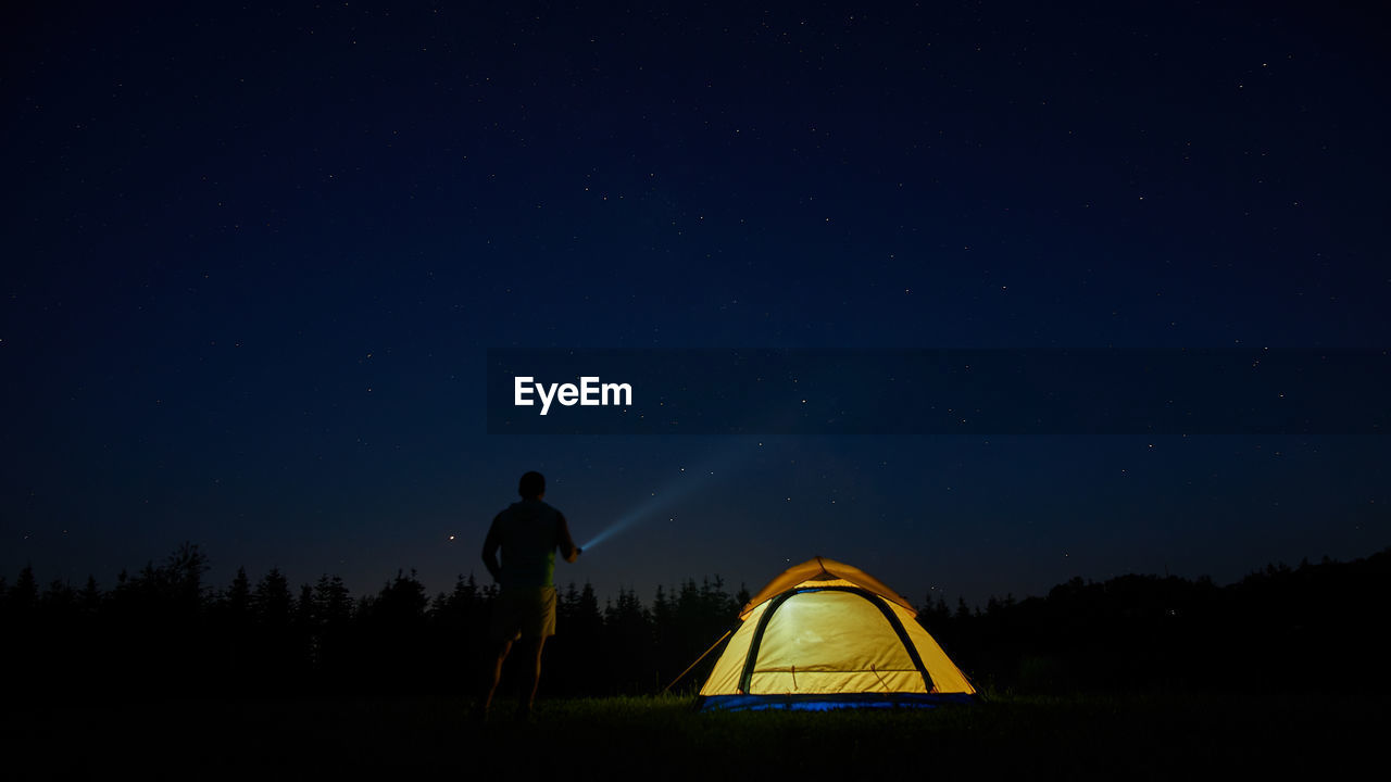 Rear view of man flashing light in sky while standing by tent at night