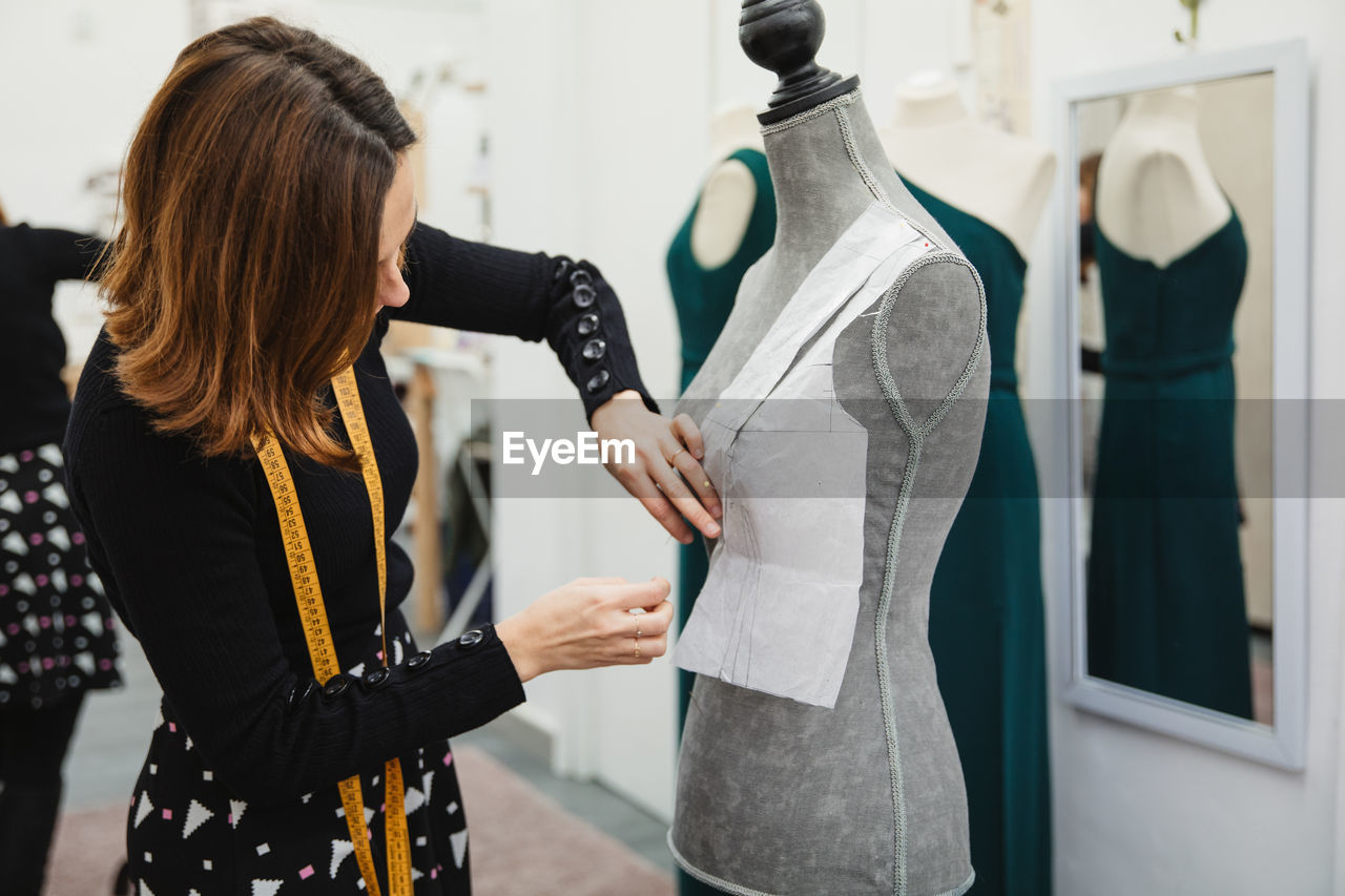 Anonymous woman with tape measure attaching paper cutout to mannequin with pin while making clothes in professional workshop