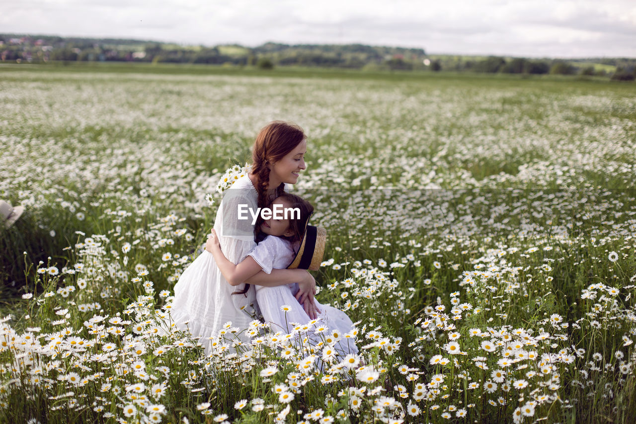 Mother with daughter in a white dress and hat stand in a daisy field in summer