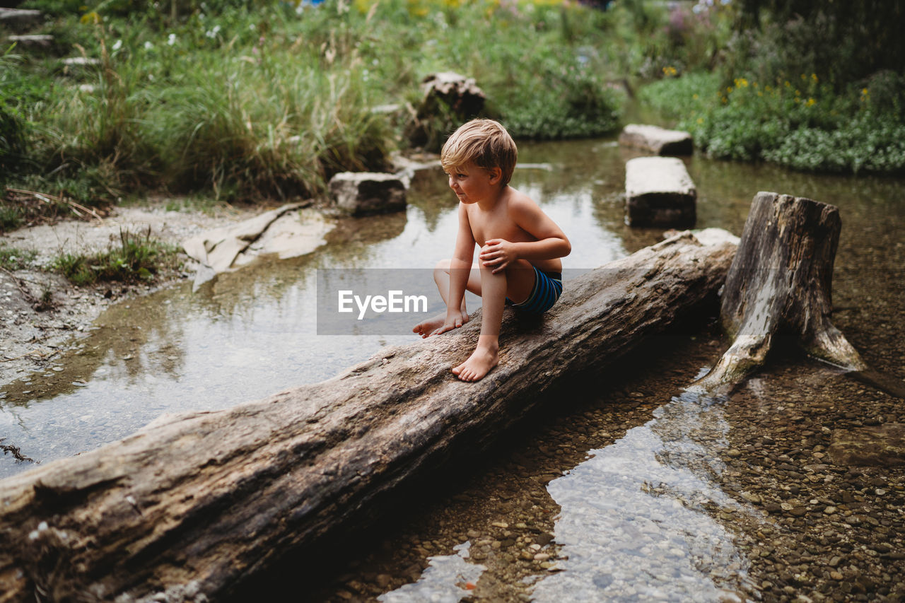 Young boy sitting on log in water in summer looking thoughtful