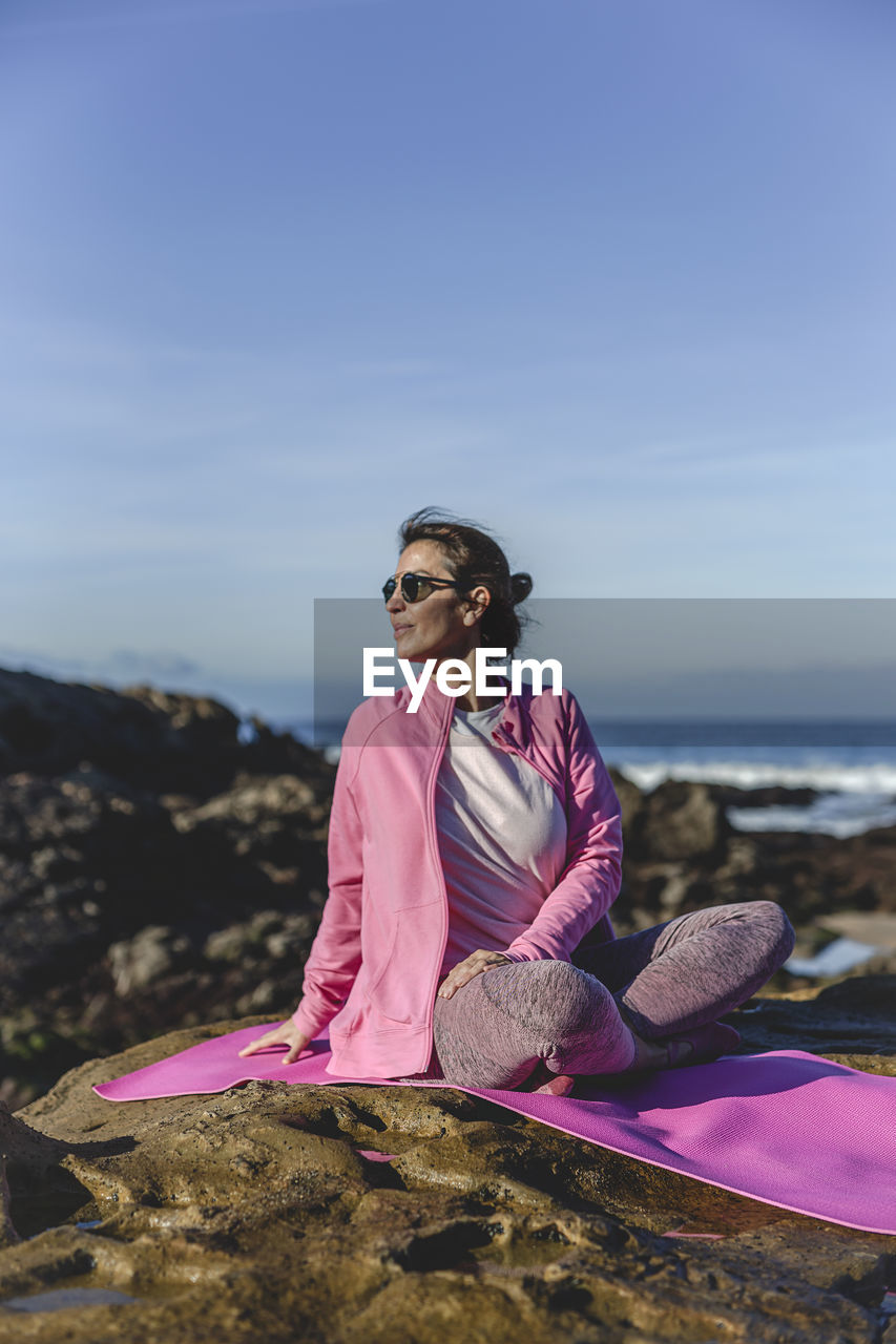 Mature woman exercising while sitting over mat on rock by sea against sky