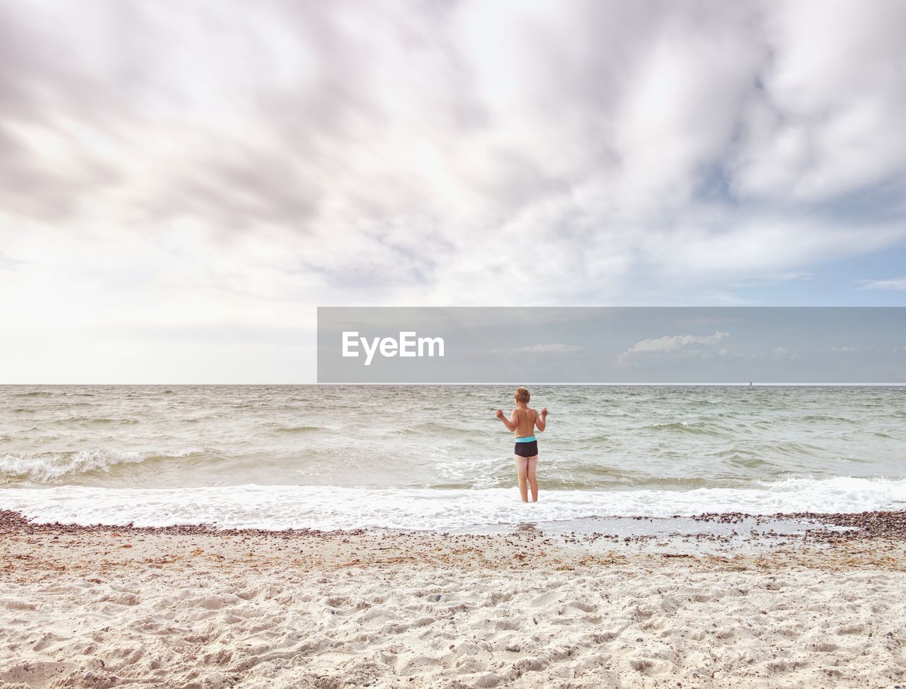 Blond hair boy stay in cold sea tide. kid on stony beach with foamy waves. windy day, cloudy sky