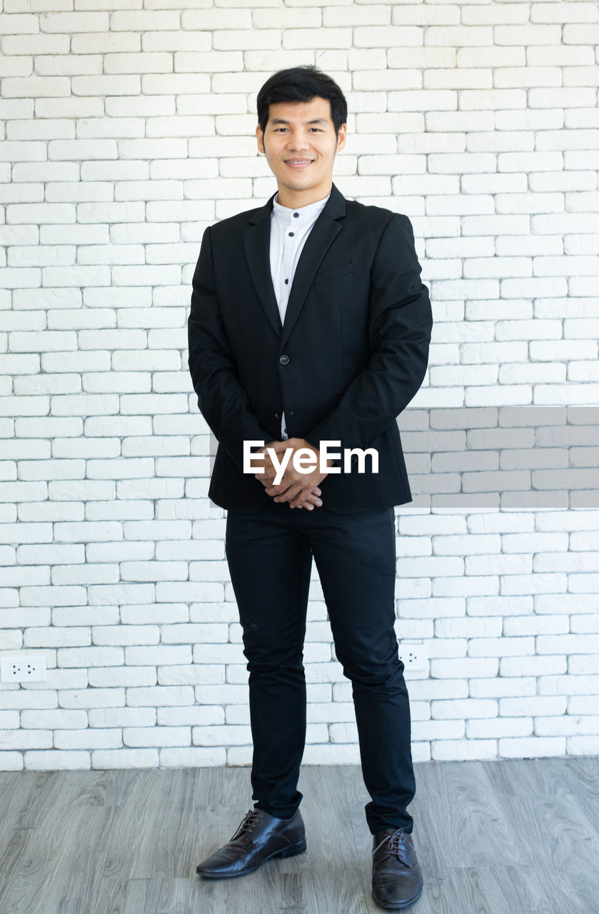 Portrait of young asian businessman in suit standing and smile with white brick background. person