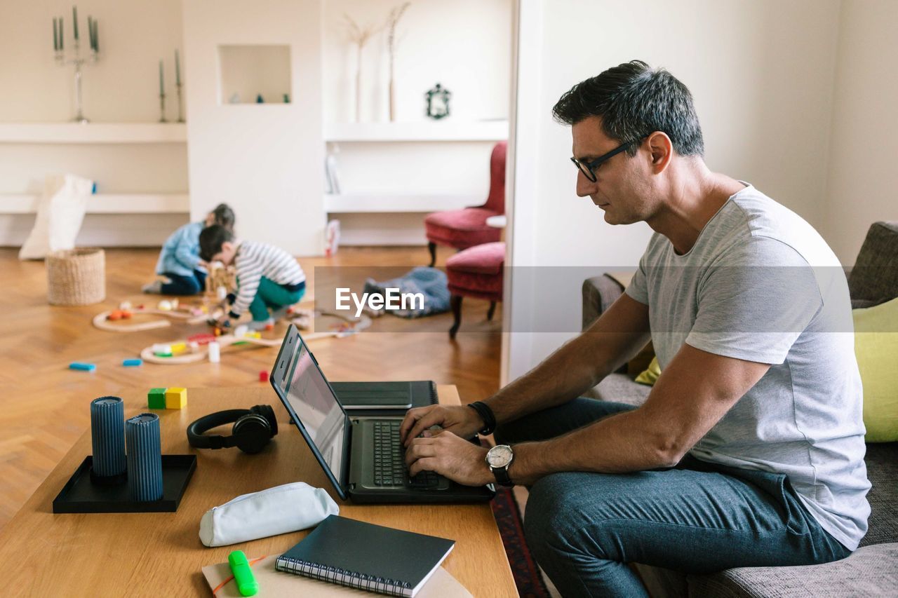 Father typing over laptop while children playing in background at home