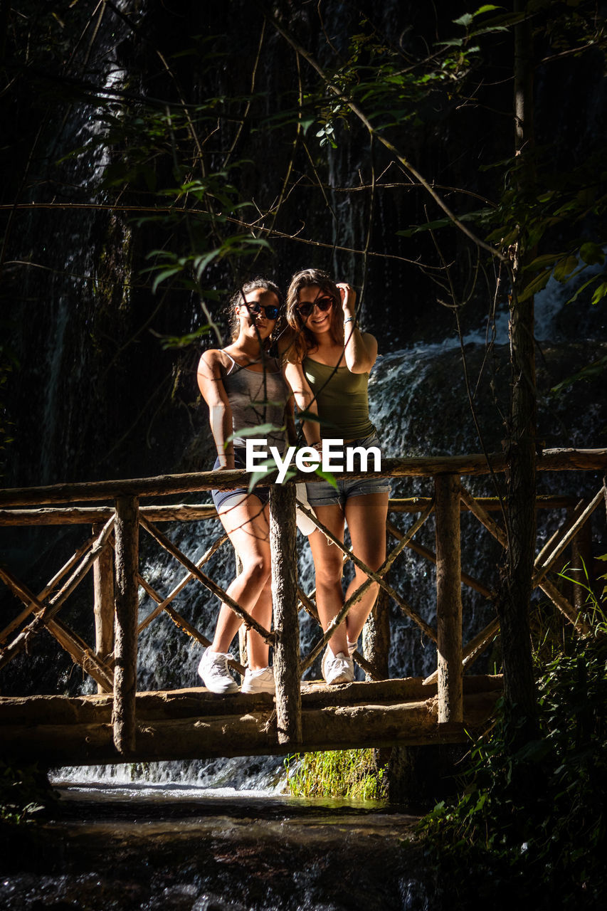 Portrait of smiling friends standing on footbridge over stream in forest