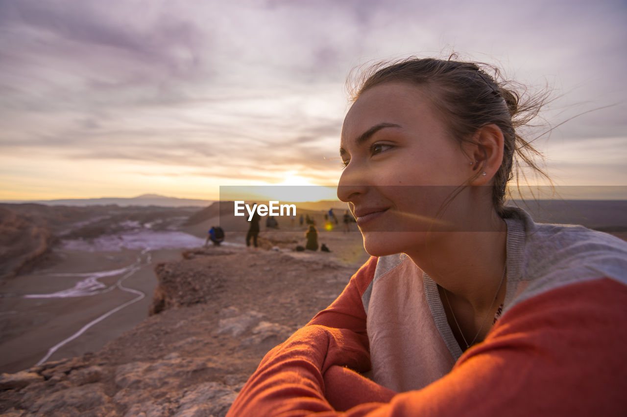 Side view of smiling young woman sitting on cliff against sky during sunset