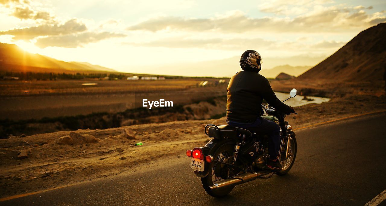 REAR VIEW OF MAN RIDING MOTORCYCLE ON LANDSCAPE AGAINST SKY