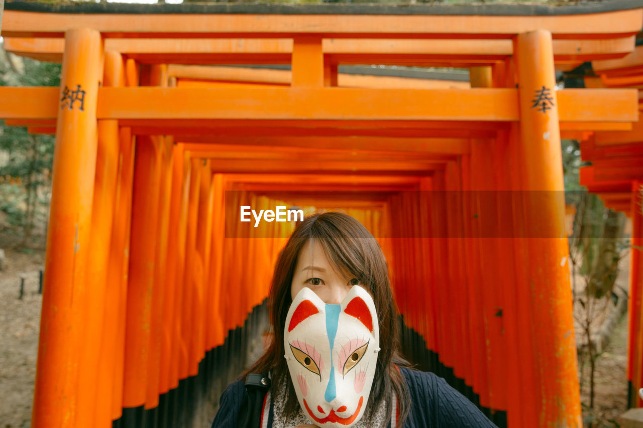 Portrait of young woman holding mask against torii gates