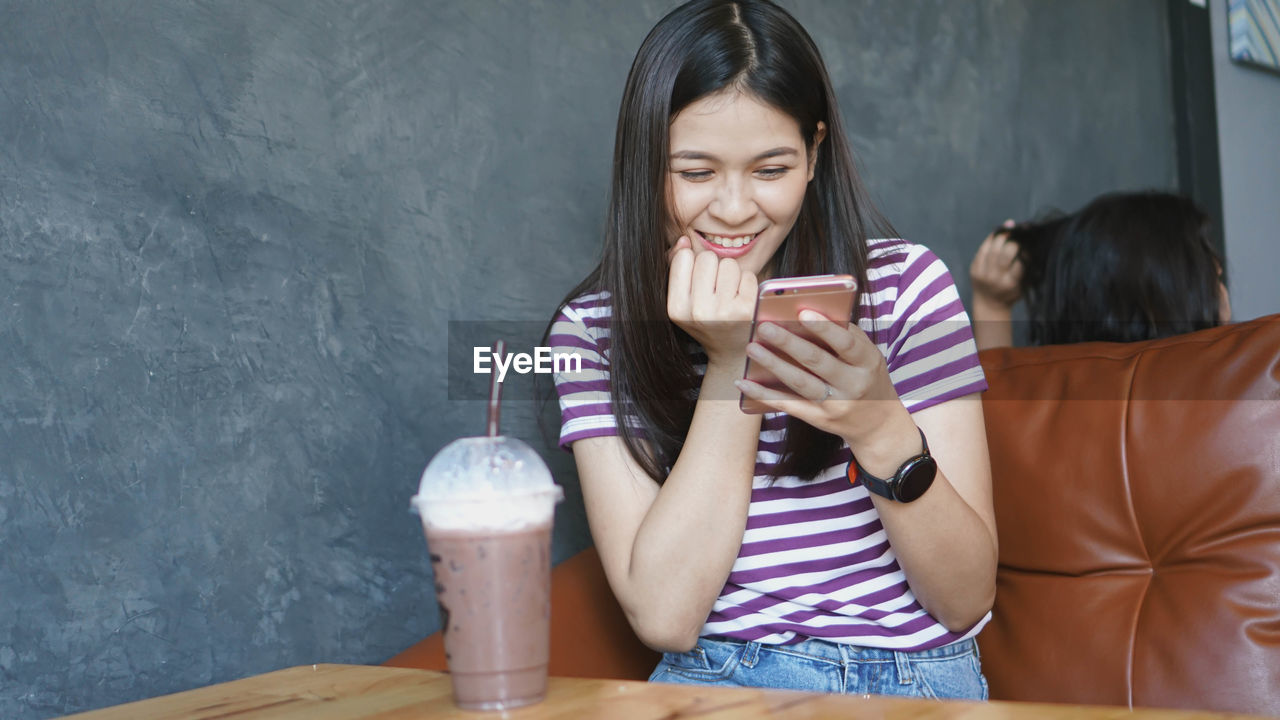 Cheerful young woman using smart phone sitting at table with coffee in cafe