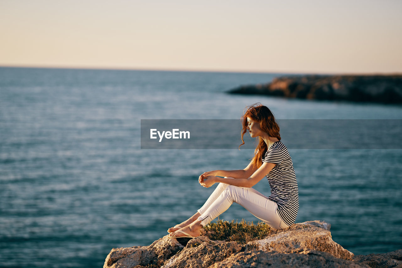 YOUNG WOMAN SITTING ON ROCK BY SEA AGAINST SKY AT SUNSET