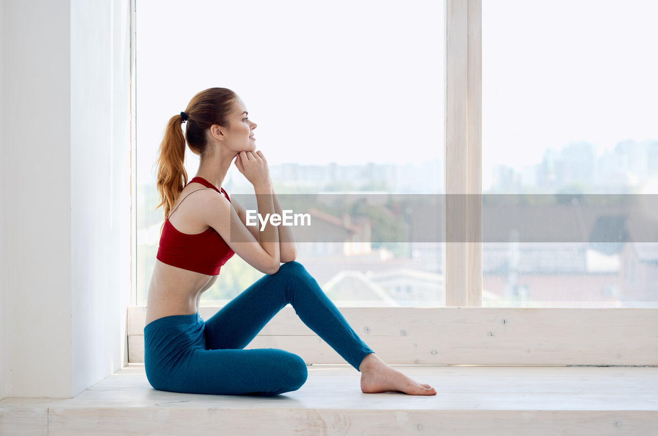 SIDE VIEW OF WOMAN SITTING ON WINDOW