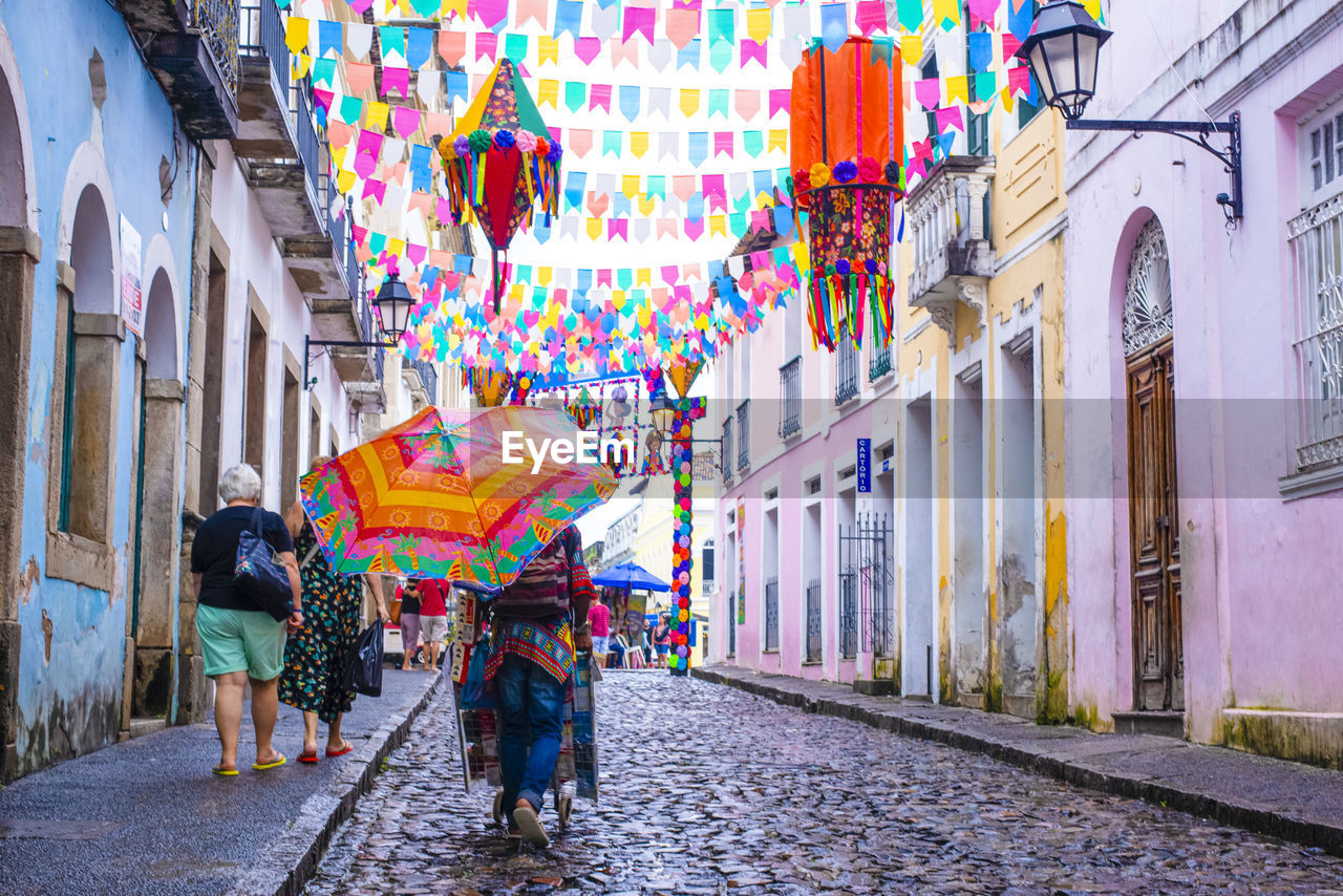 architecture, street, city, road, multi colored, building exterior, built structure, walking, adult, umbrella, women, men, infrastructure, urban area, city life, building, full length, the way forward, lifestyles, decoration, day, outdoors, city street, group of people, celebration, alley, cobblestone, town, tradition, rear view, travel destinations, travel, transportation, person, motion, nature, footpath