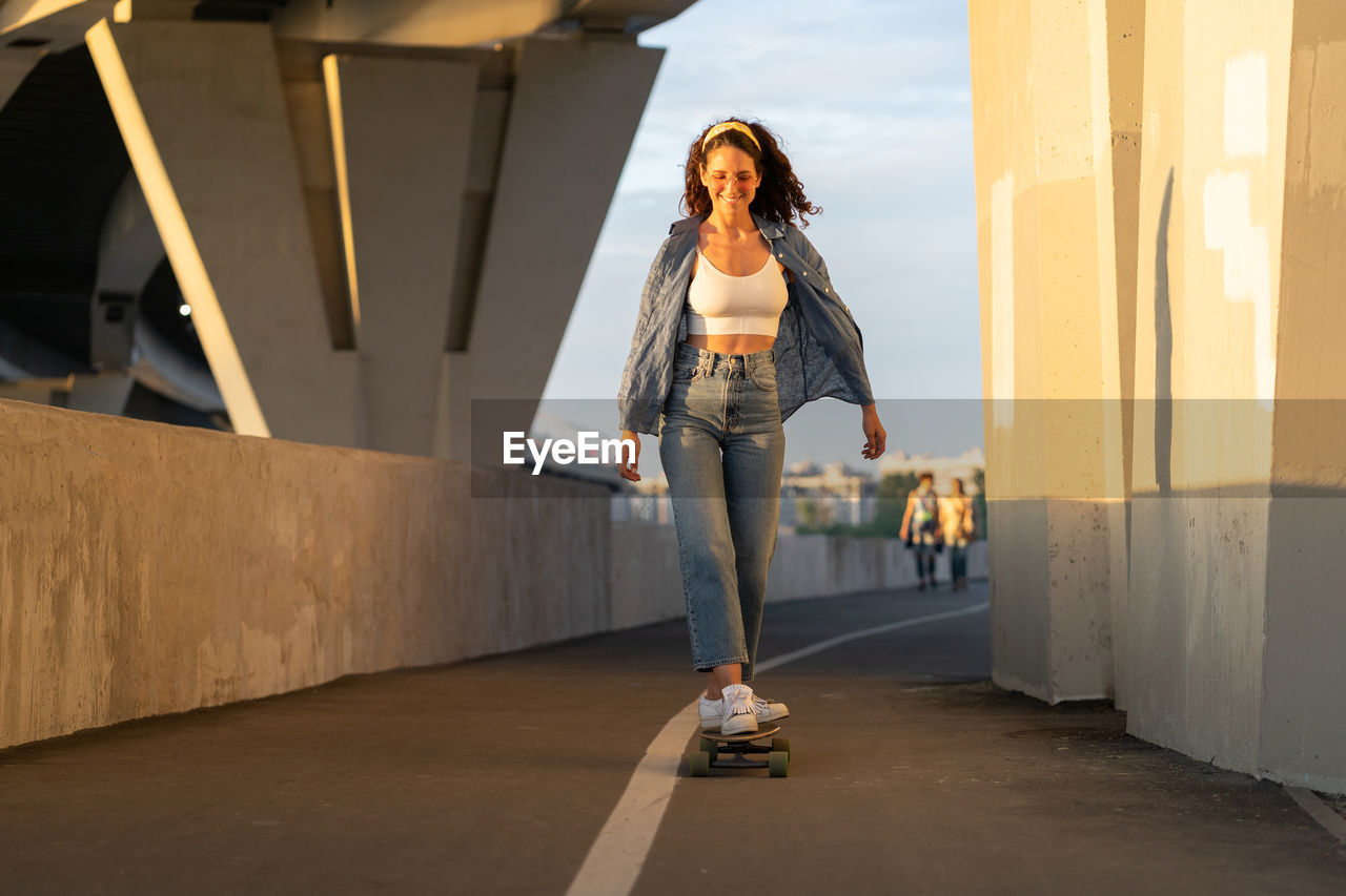 Trendy young girl skate on longboard dressed denim shirt, white top and stylish sunglasses at sunset