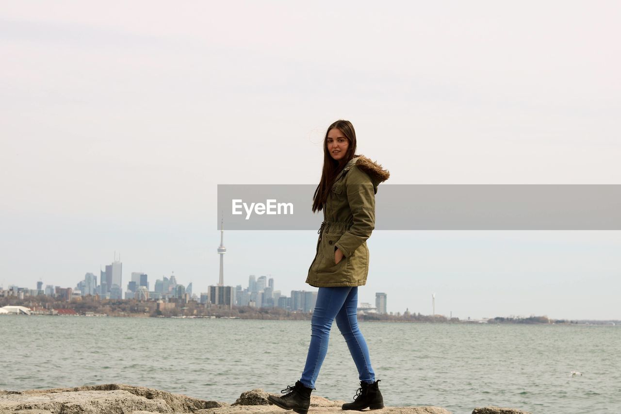 Full length portrait of young woman walking by river with cn tower in background