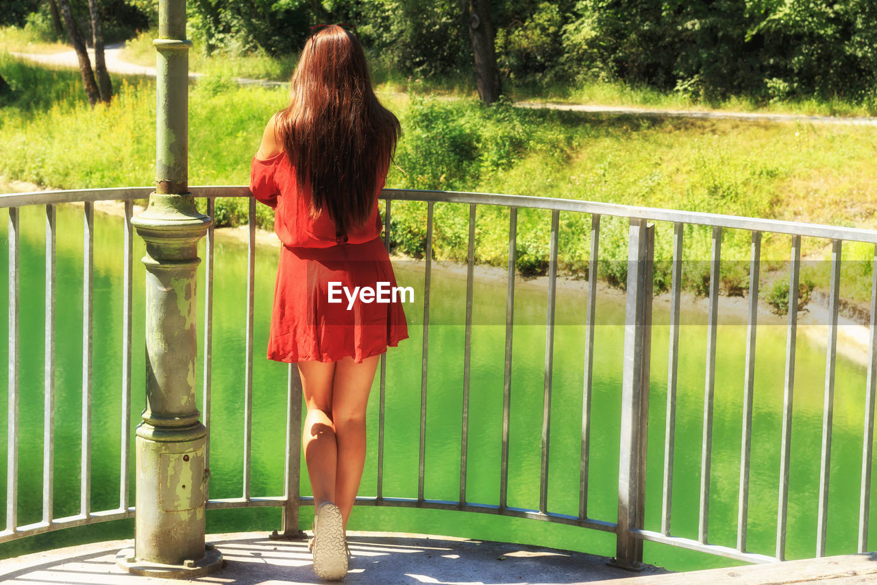 Rear view of young woman looking at lake while standing by railing during sunny day