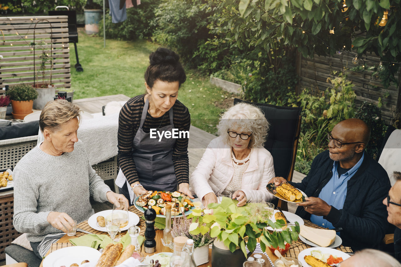 Woman serving grilled meal to senior friends sitting at dining table during dinner party in back yard