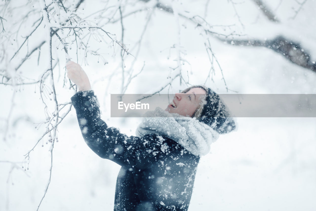 Side view of smiling woman standing by snow covered tree during winter