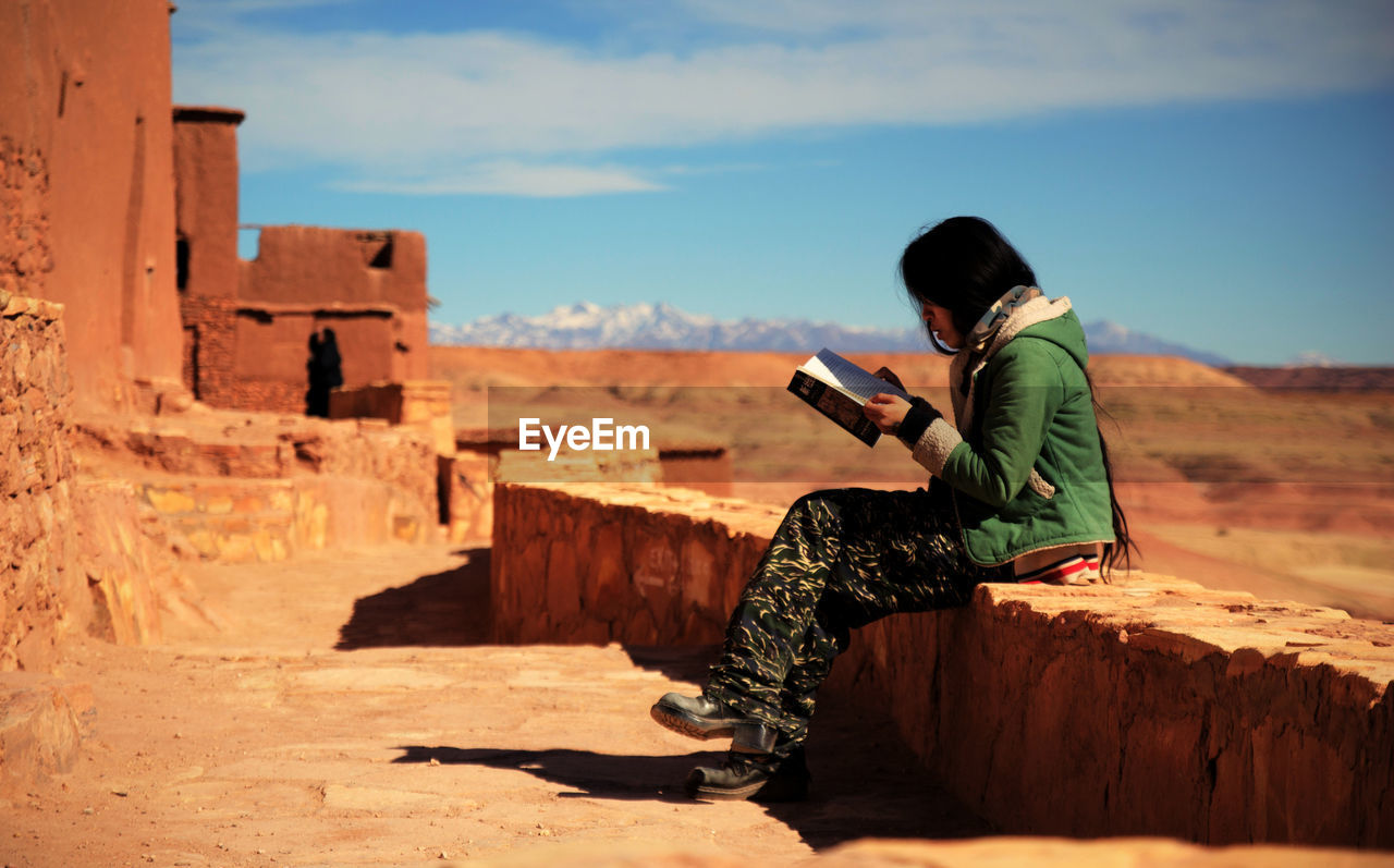 Side view of young woman reading book while sitting on retaining wall against sky