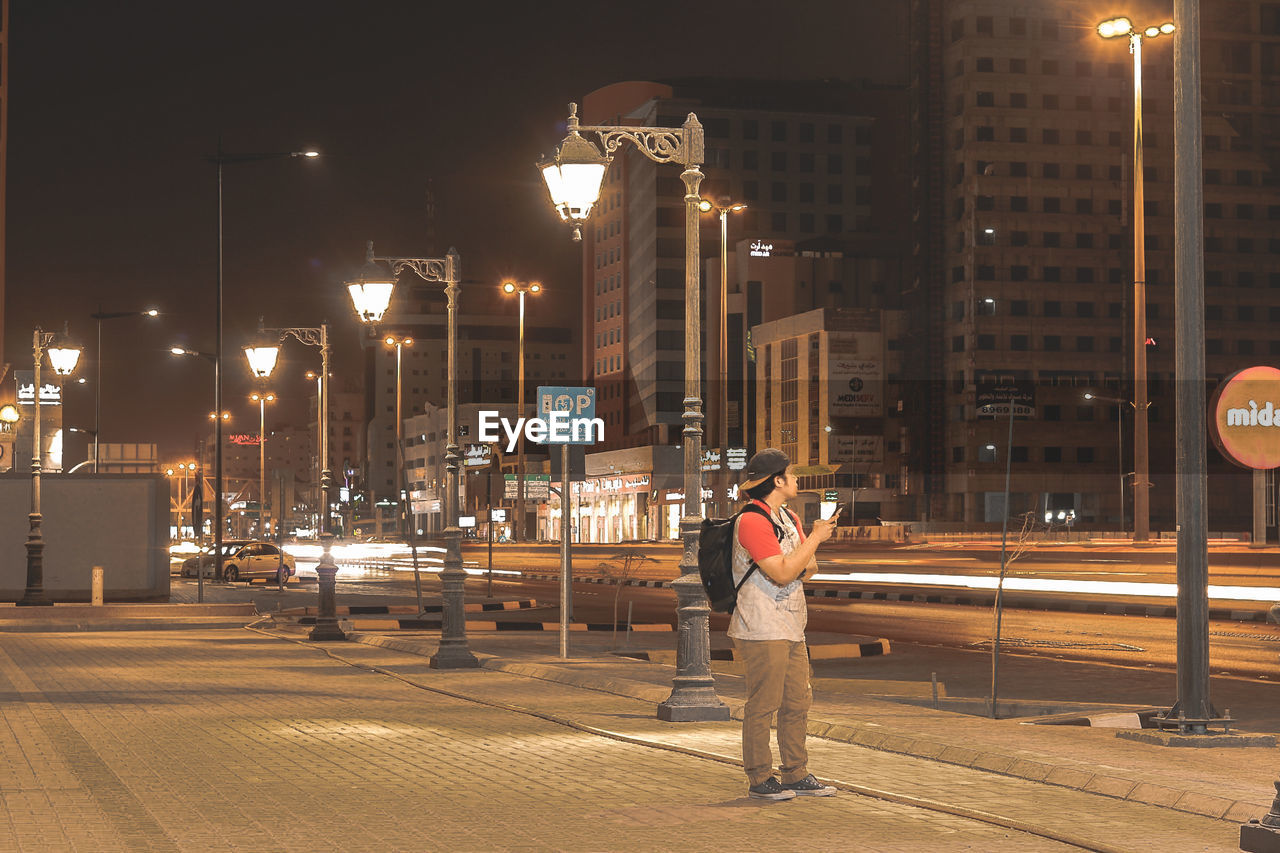 REAR VIEW OF MAN STANDING BY ILLUMINATED STREET AT NIGHT