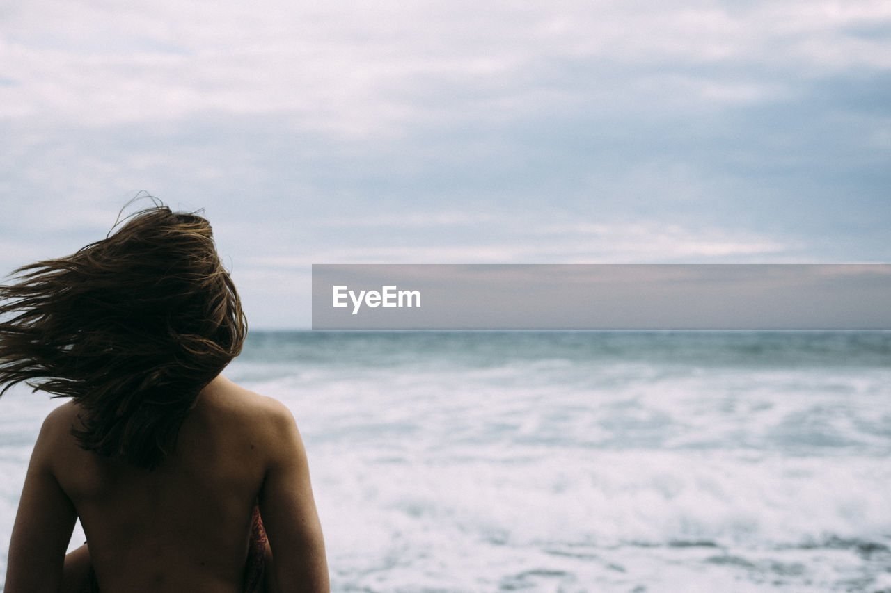 Rear view of shirtless mid adult woman standing at beach against cloudy sky