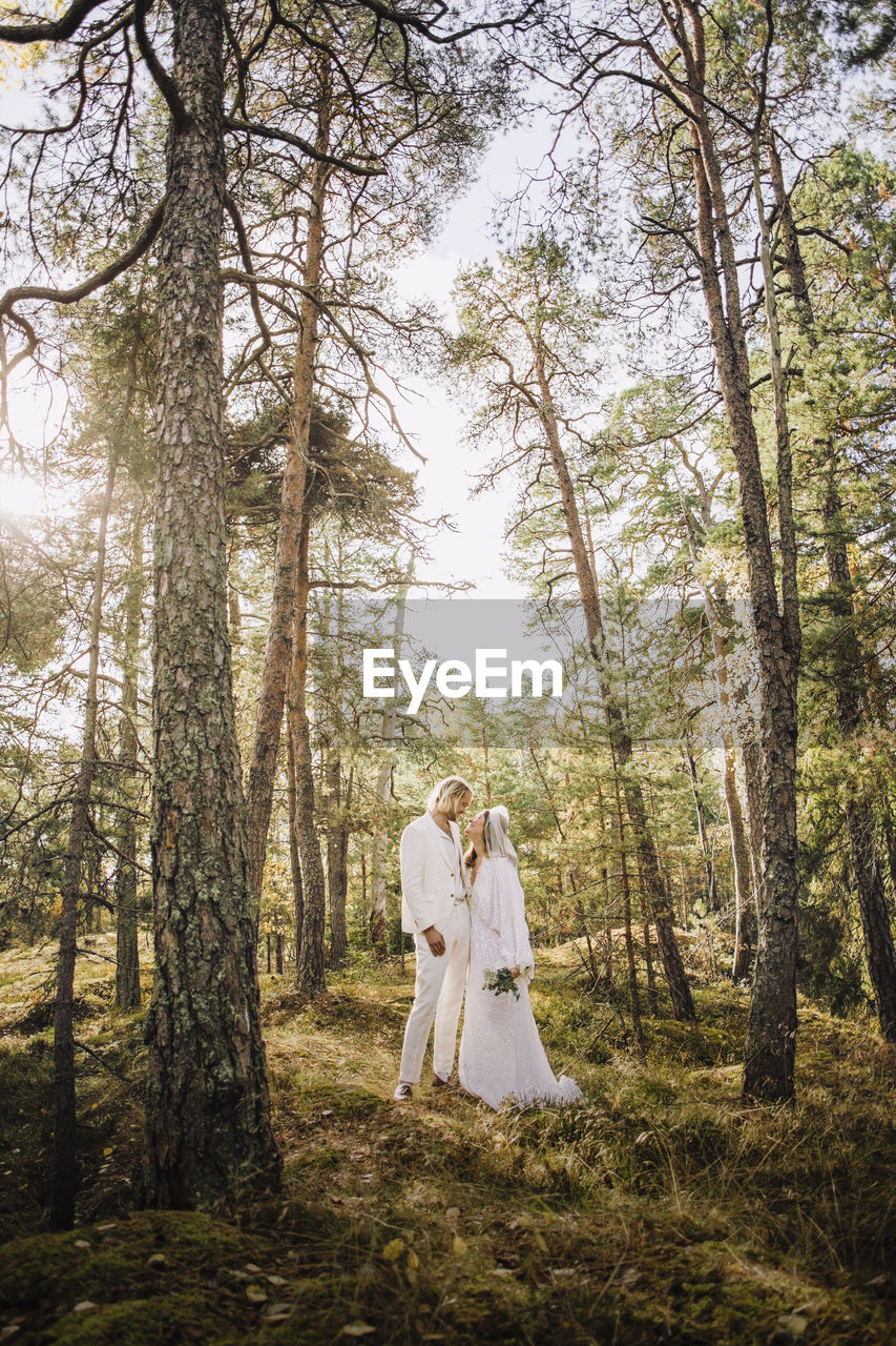 Mid adult bride holding bouquet looking at groom standing amidst trees in forest