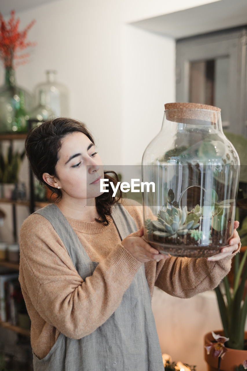Side view of female in casual clothes holding big transparent glass terrarium closed with cork lid and looking at small green succulents inside while standing against blurred interior of modern flower shop