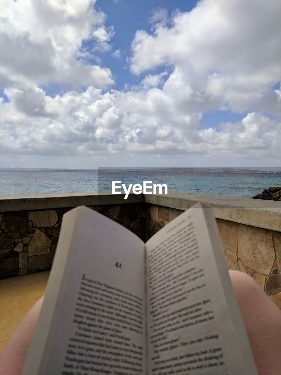 Midsection of person with book by sea against cloudy sky