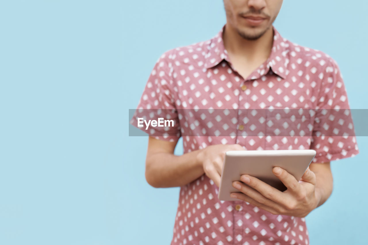 one person, studio shot, colored background, blue background, pattern, polka dot, front view, indoors, adult, blue, technology, holding, standing, digital tablet, copy space, waist up, wireless technology, casual clothing, young adult, communication, spotted, pink, portrait, looking, button down shirt, internet, person, men, computer network, computer, smiling, emotion, portability