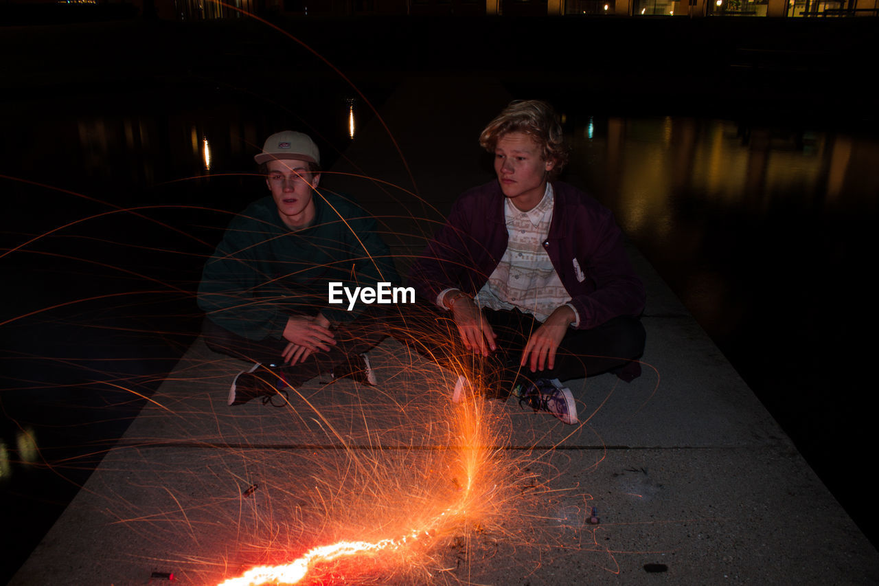 Young men sitting in front of burning firework over lake at night