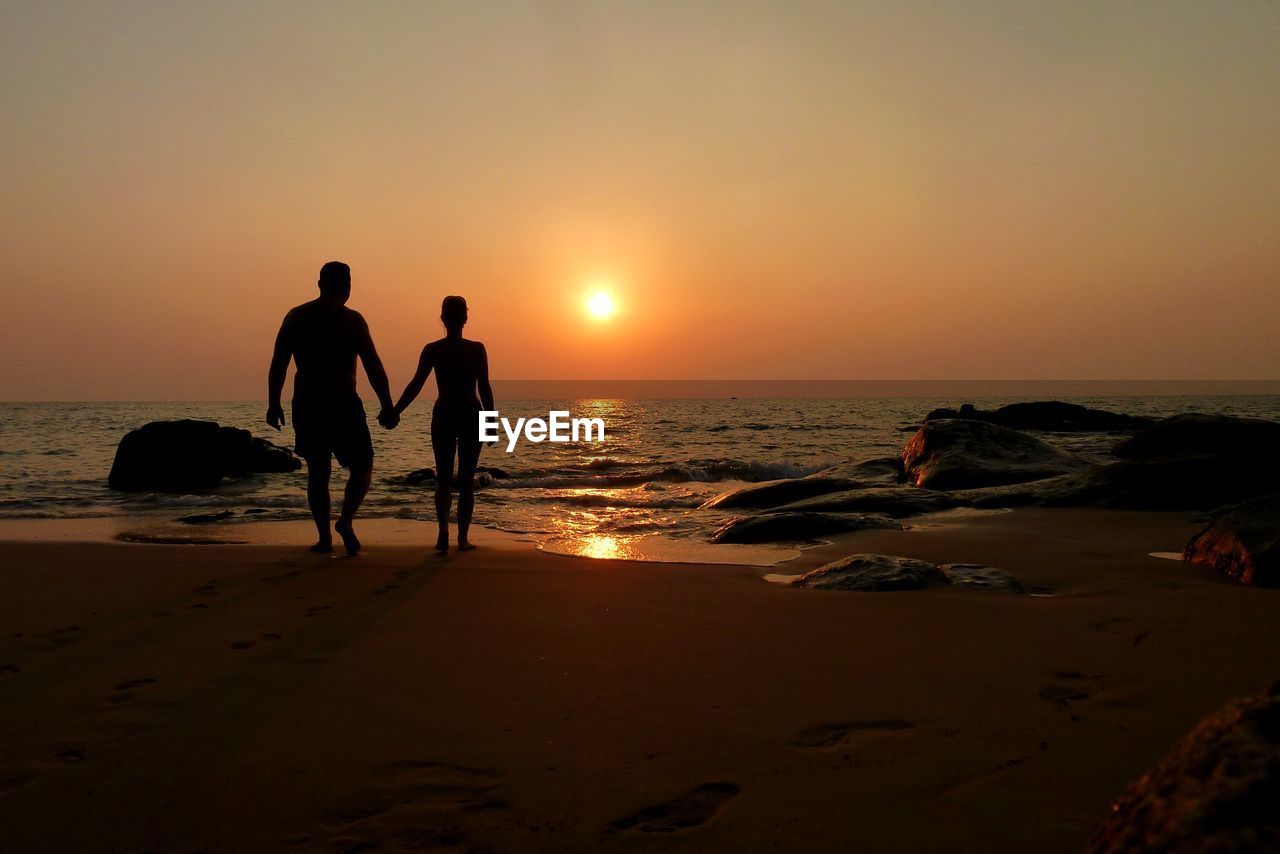 Rear view of silhouette man and woman holding hands while walking on shore during sunset