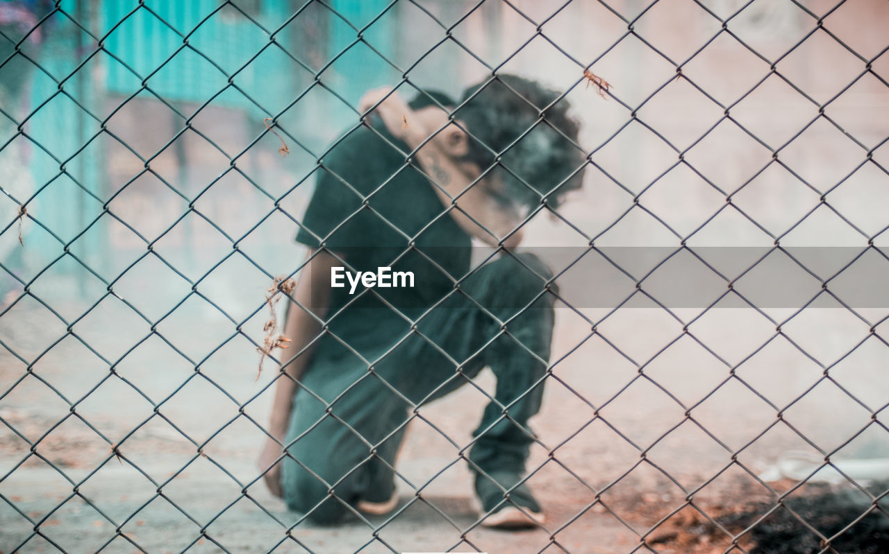 Full length of man kneeling on footpath seen through chainlink fence