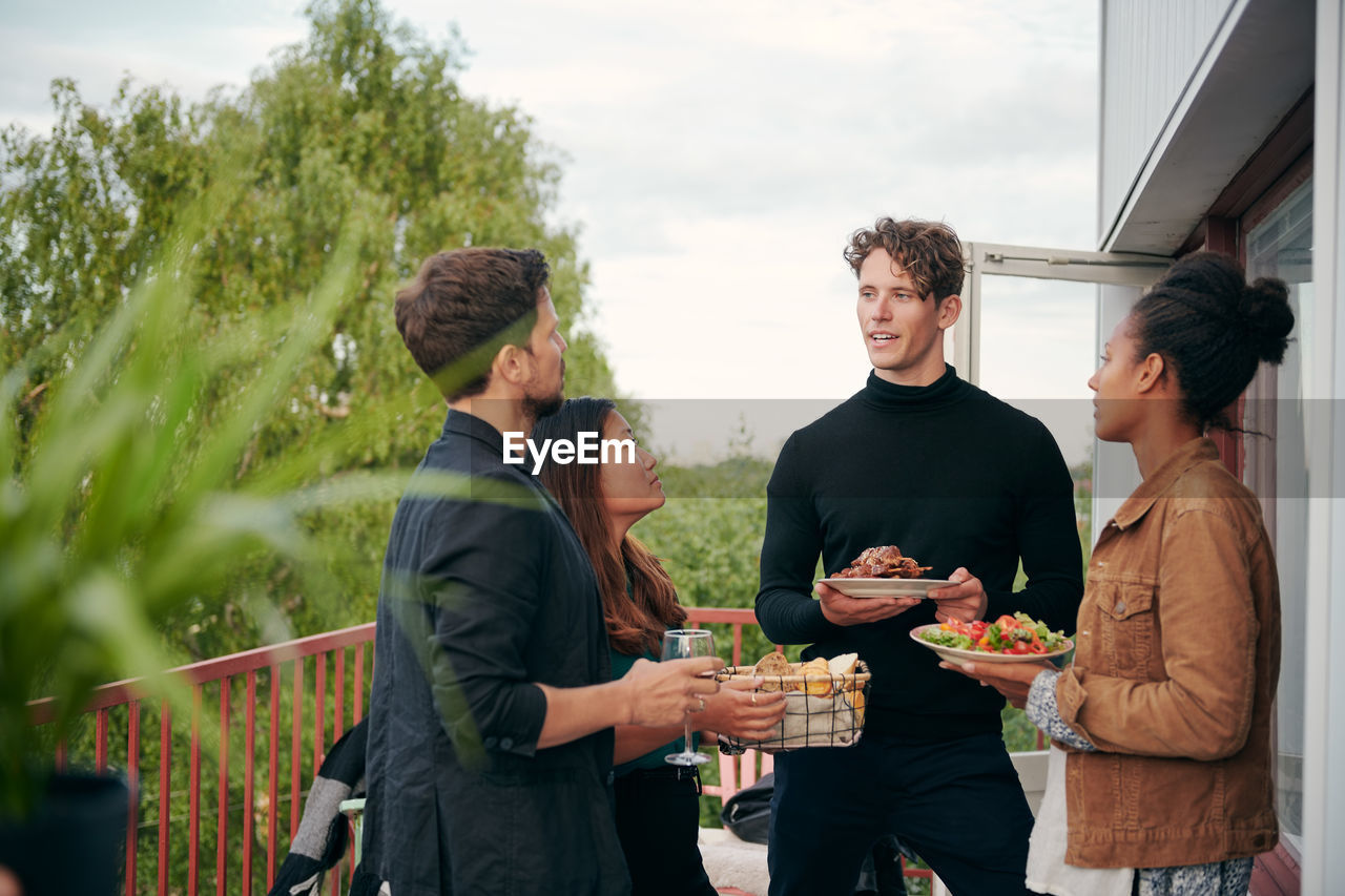 Young man talking with friends while holding meal during party on terrace