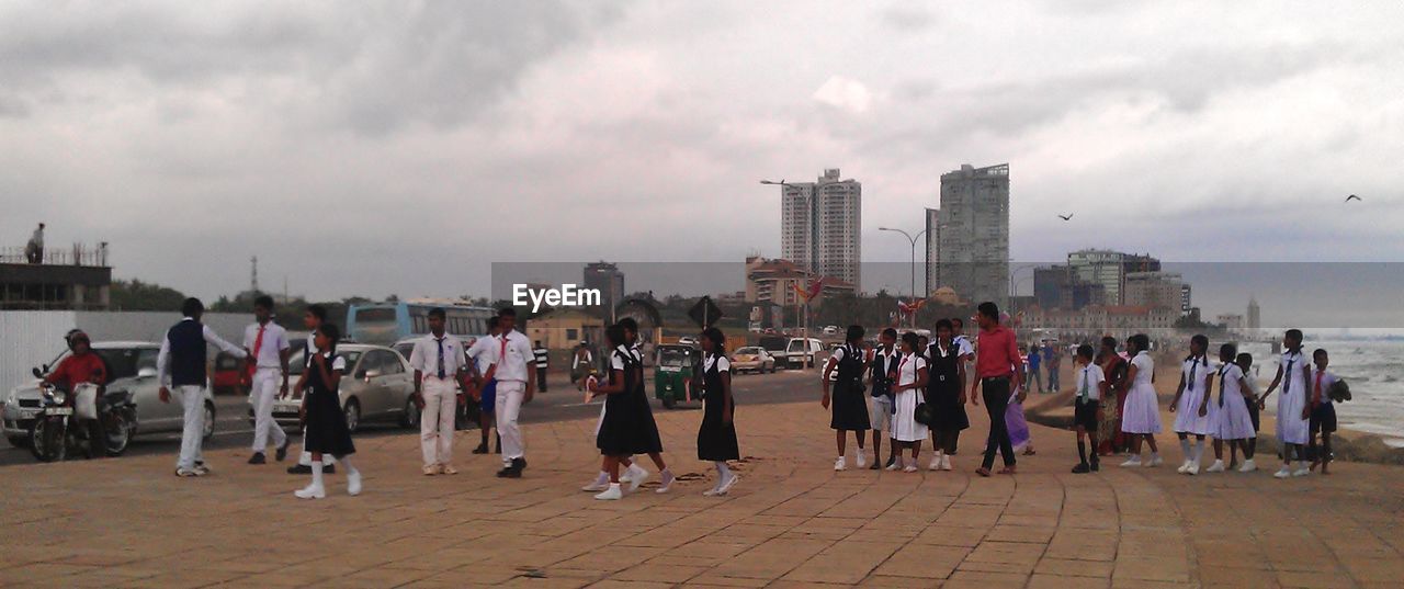 PEOPLE WALKING ON PROMENADE AGAINST CLOUDY SKY