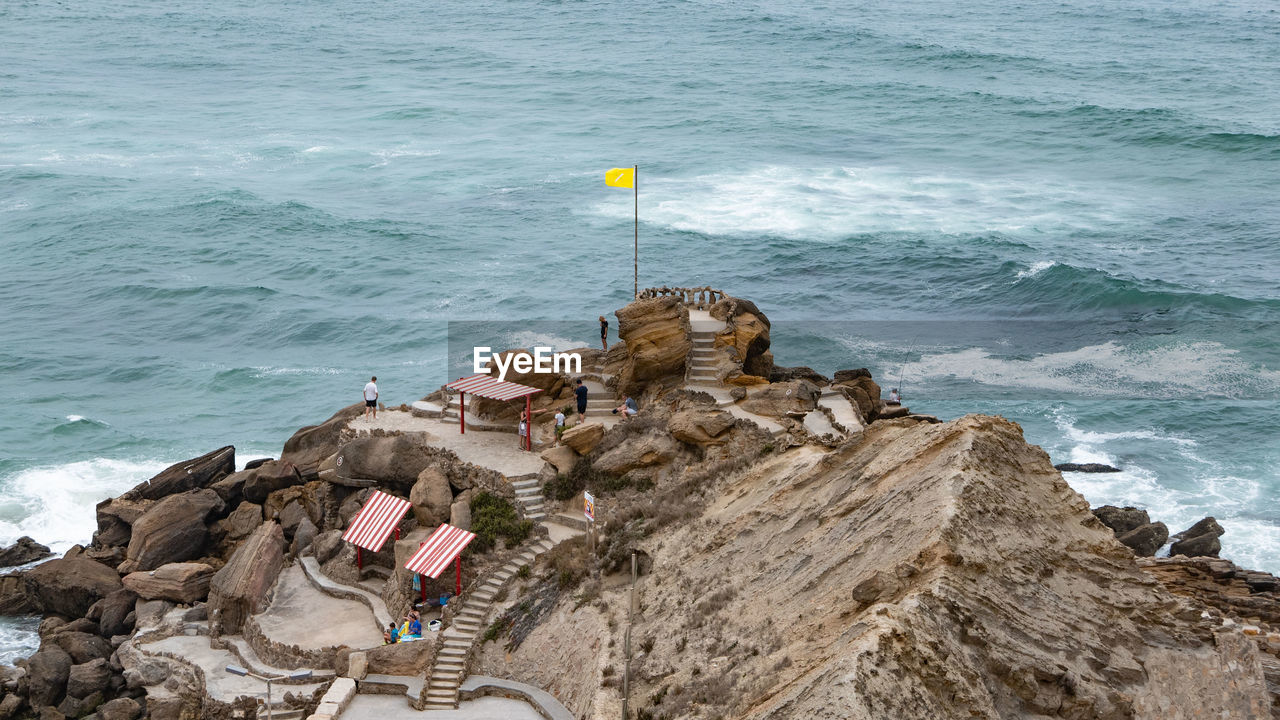 sea, water, coast, terrain, rock, ocean, cliff, land, shore, nature, beach, tower, high angle view, wave, day, no people, motion, outdoors, beauty in nature, breakwater, architecture, scenics - nature, rock formation, flag, military