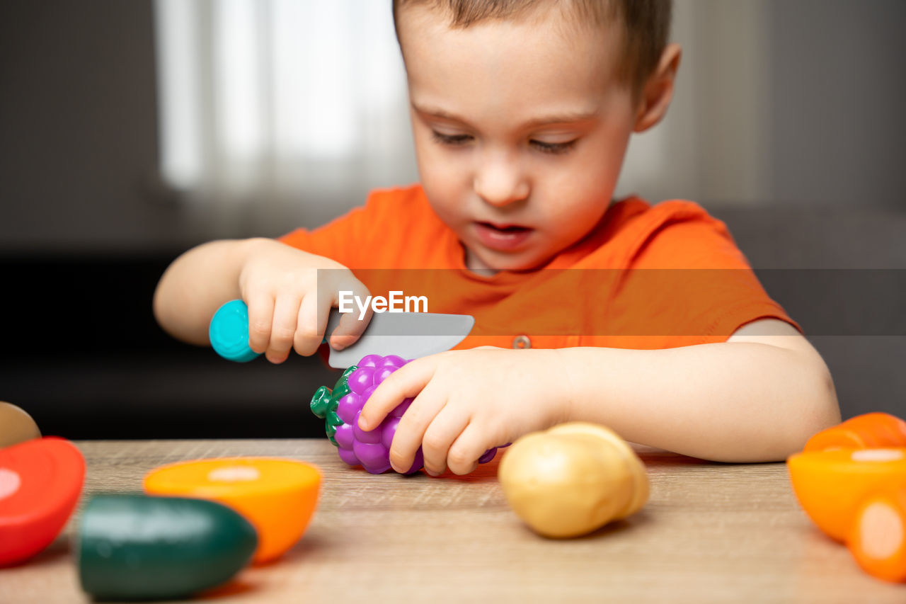 A cute 3 year toddler boy plays with toy fruits or vegetables and sitting at the table.